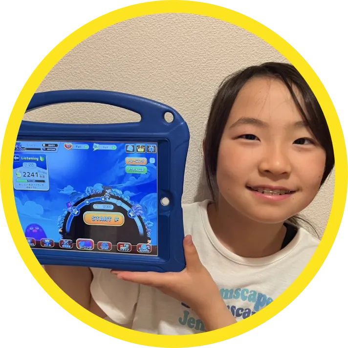 タブレットで英語学習アプリを開きながら笑顔を見せる小学生の女の子。アプリの画面にはリスニングスコアやスタートボタンが表示されている。